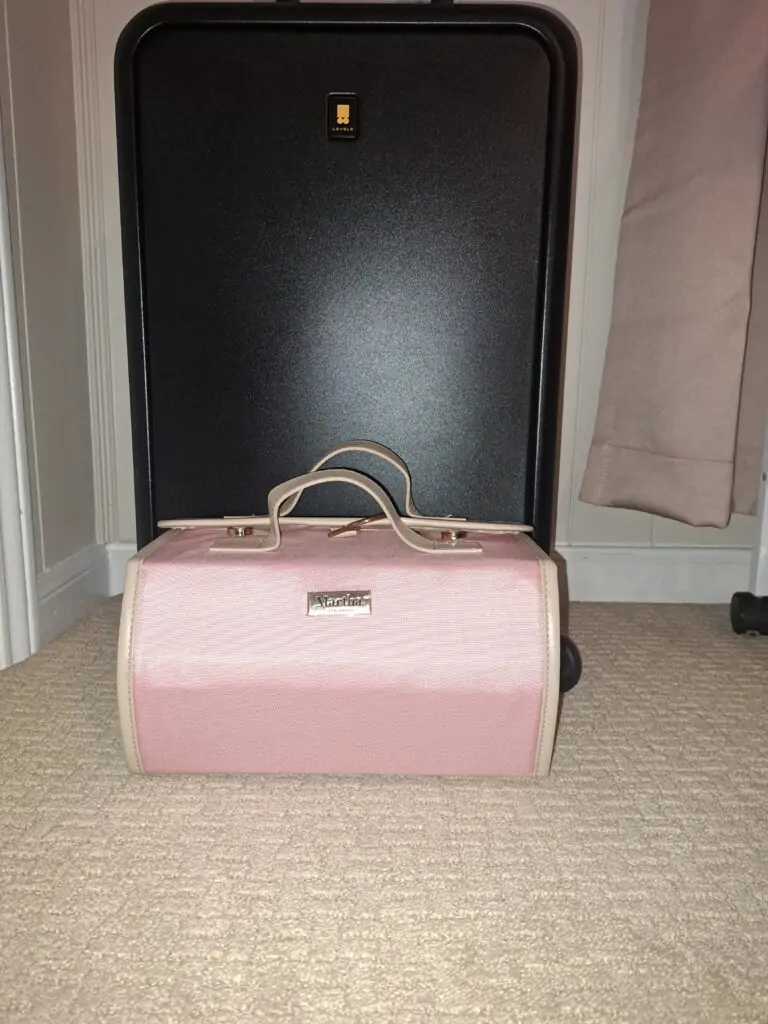 A closed roll-up travel organizer with a structured pink and beige design, branded with the name "Martha" on a silver plate. It’s neatly placed upright in front of a black suitcase on a beige carpeted floor.