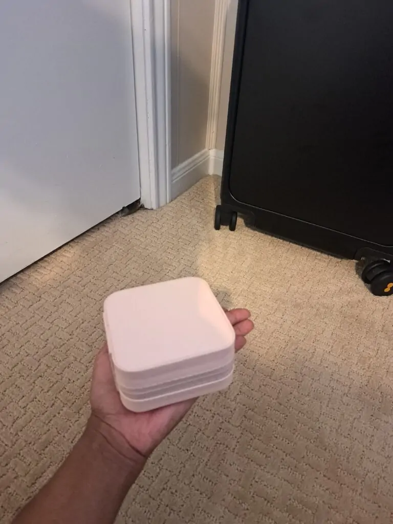 A person holds a compact, square-shaped blush pink travel jewelry case with a soft matte finish. The case is shown closed, resting in the palm of their hand, with a black suitcase and beige carpeted floor in the background.