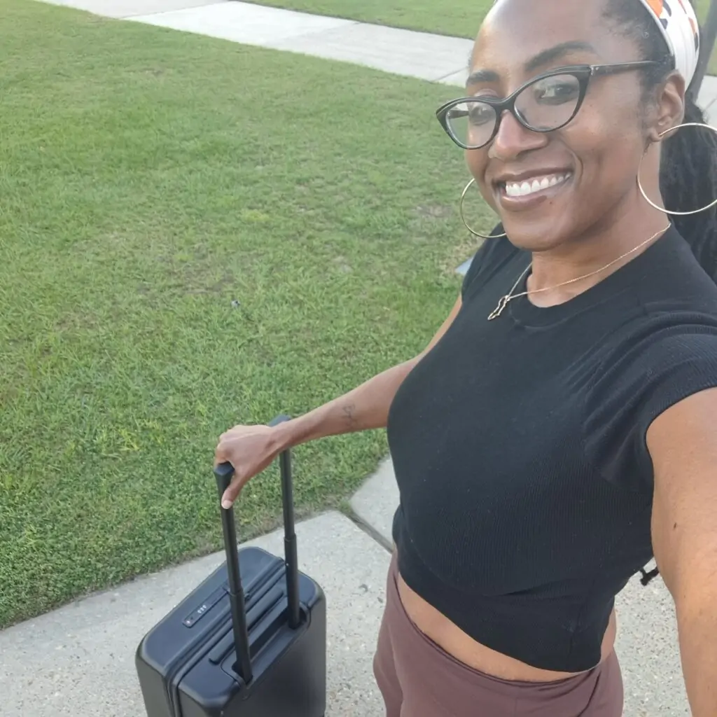 A smiling woman takes a selfie outdoors while pulling a sleek black carry-on suitcase. She’s wearing a fitted black crop top, high-waisted mauve pants, hoop earrings, and a gold necklace with an Africa-shaped pendant, with her hair wrapped in a printed headscarf. She stands on a sidewalk next to a grassy lawn.
