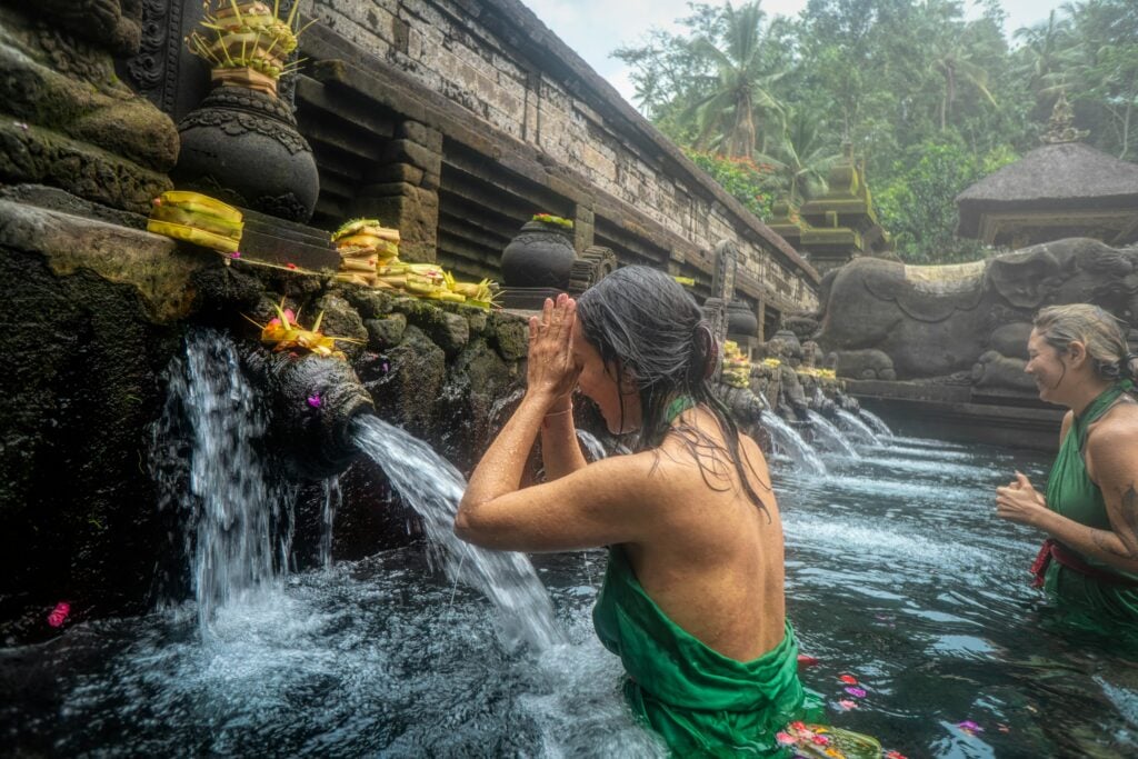 Two women dressed in green sarongs participate in a ritual purification at a Balinese water temple, standing beneath stone spouts flowing with spring water. One woman prays with hands pressed together under the stream, while flower offerings rest atop the fountain ledge above.