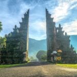 A traditional Balinese temple gate, known as a candi bentar, stands tall and split in two against a backdrop of misty mountains and a glowing morning sky. Lush greenery surrounds the ornate stone structure, with ceremonial umbrellas and fabric decorations at the base of each side of the gate.