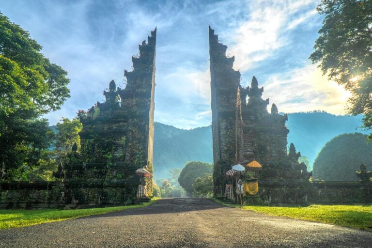 A traditional Balinese temple gate, known as a candi bentar, stands tall and split in two against a backdrop of misty mountains and a glowing morning sky. Lush greenery surrounds the ornate stone structure, with ceremonial umbrellas and fabric decorations at the base of each side of the gate.