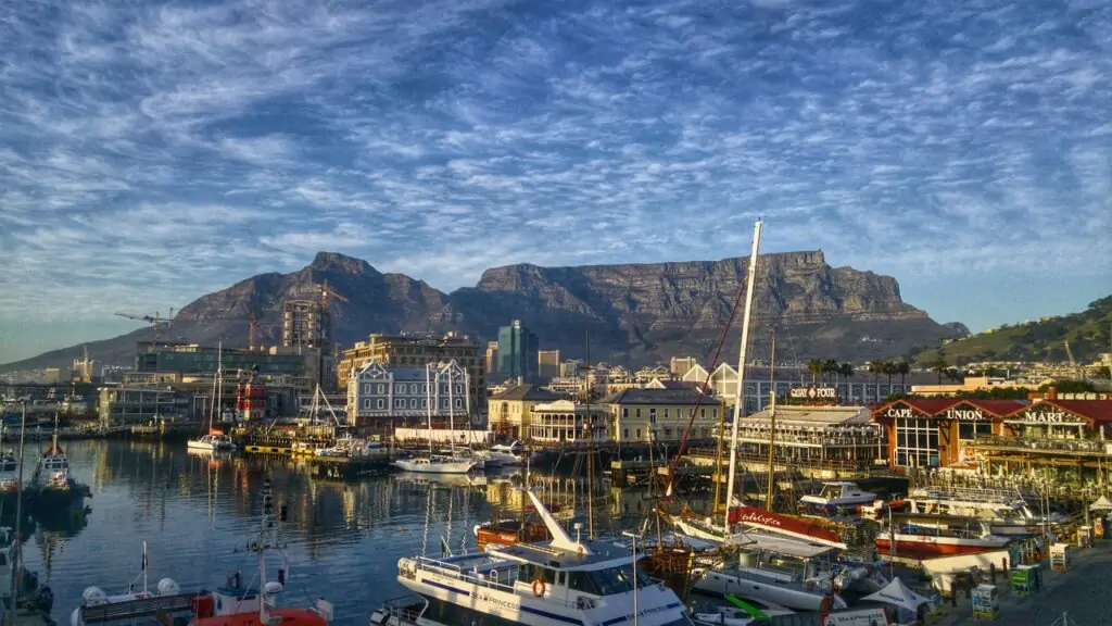 A scenic view of Cape Town’s V&A Waterfront with boats docked in the marina, colorful buildings, and Table Mountain rising prominently in the background under a partly cloudy sky. The lively harbor area includes signs for shops and restaurants like "Cape Union Mart" and "Quay Four."