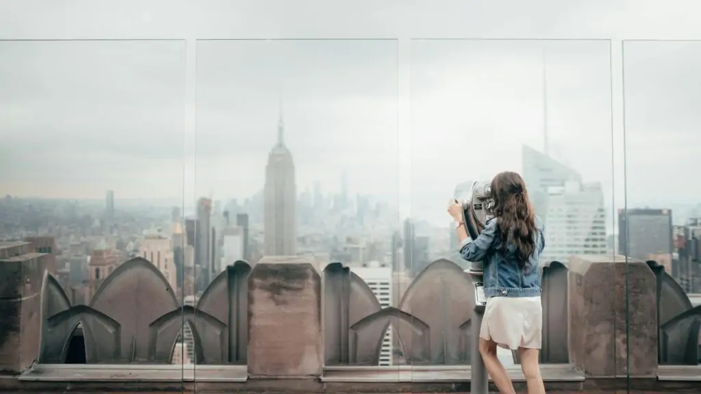 A woman in a white dress and denim jacket looks through a coin-operated viewer on a rooftop observation deck, gazing at the New York City skyline. The Empire State Building rises prominently in the misty background, surrounded by other skyscrapers under an overcast sky.
