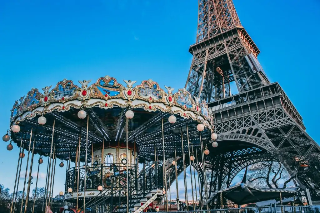 A colorful vintage carousel with ornate details and hanging globe lights sits in front of the Eiffel Tower in Paris, France. The iconic iron structure rises majestically into a clear blue sky, creating a whimsical contrast between classic amusement and architectural grandeur.