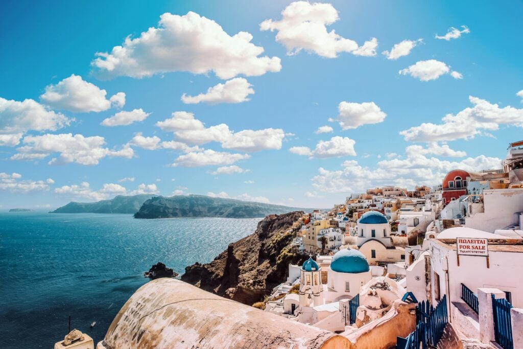 A stunning coastal view of Santorini, Greece, featuring whitewashed buildings with iconic blue domes perched on a cliffside overlooking the deep blue Aegean Sea. Fluffy clouds float across a bright blue sky, and a "For Sale" sign in Greek and English is visible on one of the buildings in the foreground.