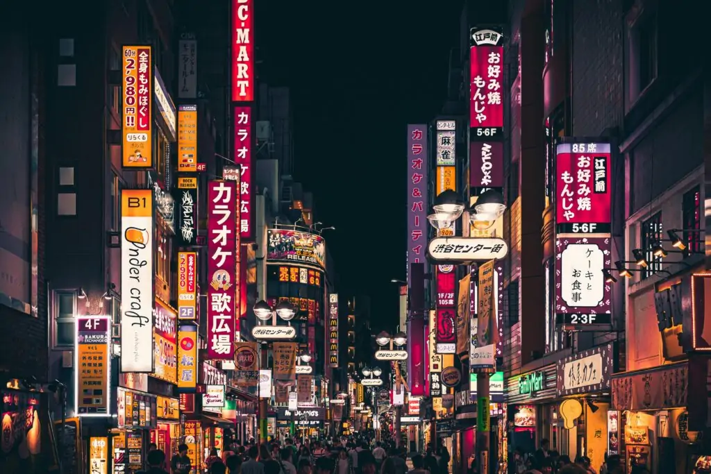 A vibrant Tokyo street scene at night, filled with colorful neon signs in Japanese advertising karaoke bars, restaurants, and shops. The street is bustling with people, and the glow of lights reflects off the buildings, creating an energetic and lively urban atmosphere.