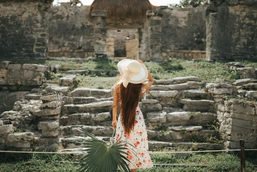 A woman in a floral dress and wide-brimmed hat stands with her back to the camera, gazing at ancient stone ruins surrounded by grass and weathered stone steps. She holds a large palm frond, adding a tropical touch to the historical site, likely located in Tulum, Mexico.