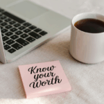 A light pink sticky note with bold black cursive text reading “Know your Worth” rests beside a laptop keyboard and a steaming white mug of coffee on a soft beige surface. The scene evokes a calm, motivational workspace vibe.